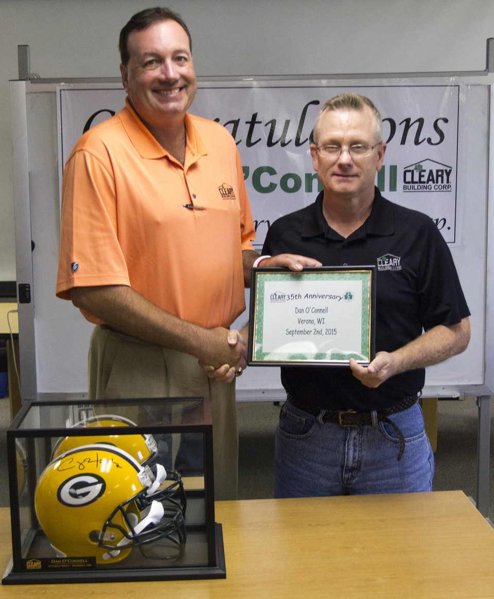 Dan is presented with an anniversary certificate and shakes hands with Sean Cleary. Packers helmet sits on table in front.