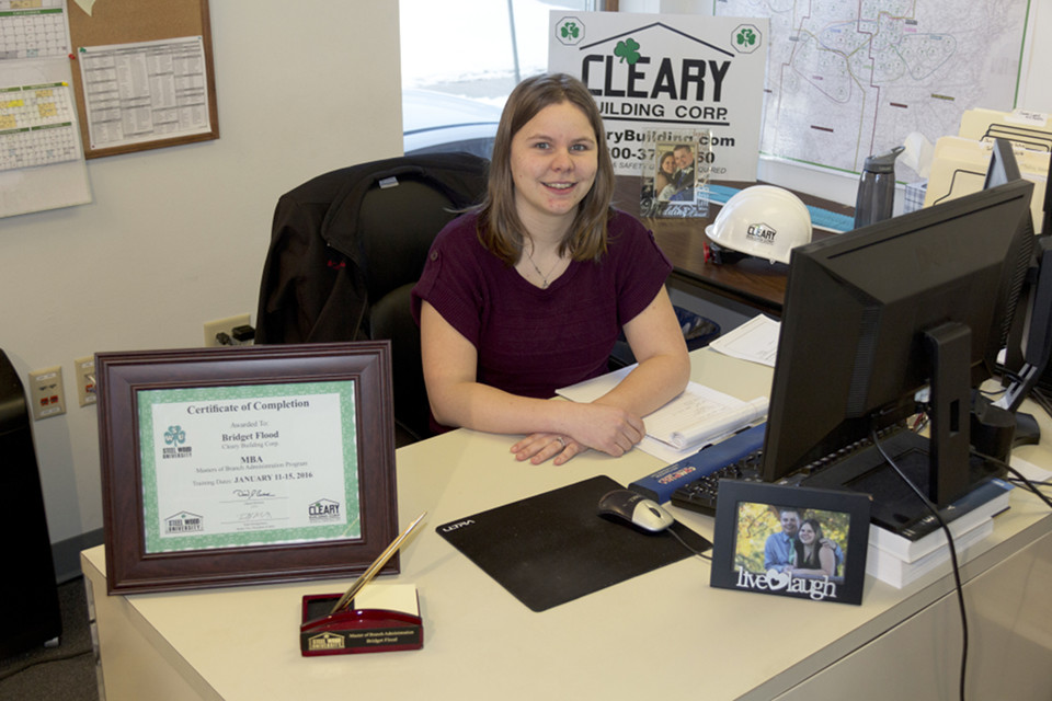 Bridge sits behind her desk which displays a Cleary Building Corp sign and certificate of completion.