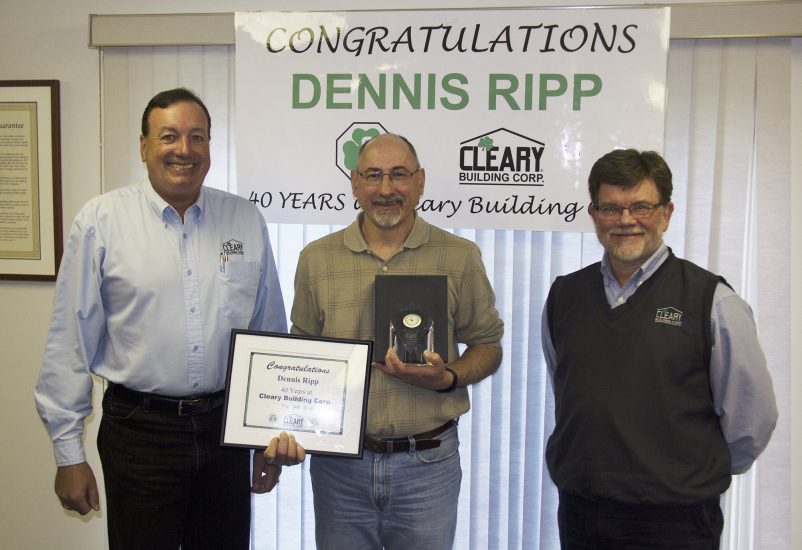 Three men smile at the Camera. Dennis (middle) holds a clock and anniversary certificate.
