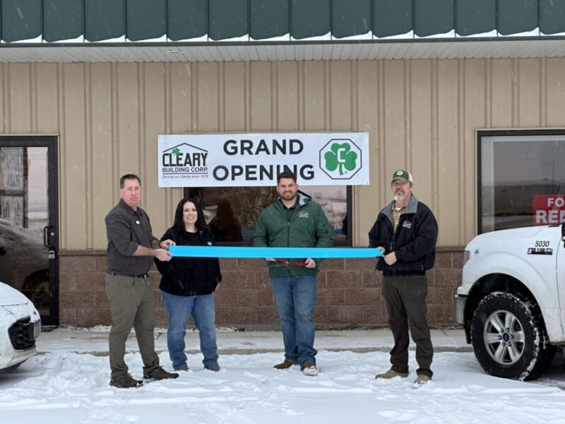 Four people standing in front of a "Grand Opening Sign," about to cut a blue ribbon.