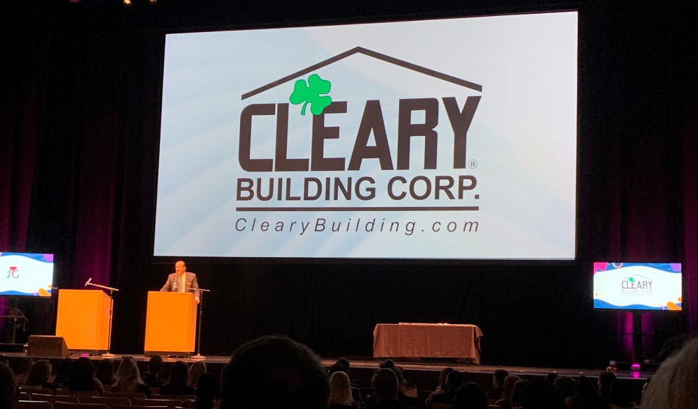 Sean Cleary stands behind a podium in the forefront. In the background is a giant screen showing the Cleary logo.